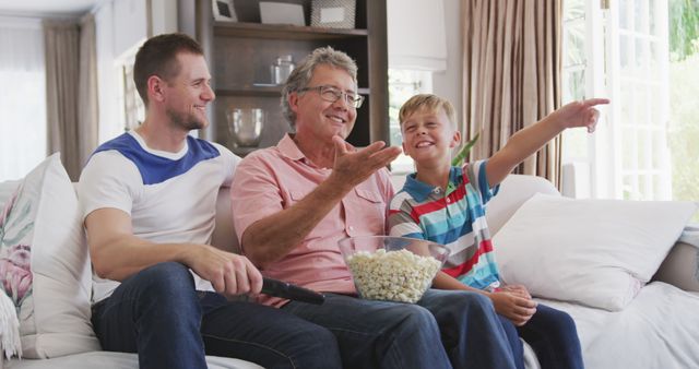 Three generations of men - a grandfather, father, and son - enjoying leisure time together on a couch at home. They are smiling and engaging in a living room filled with natural light. A bowl of popcorn is centered on the father's lap, suggesting a movie or TV show viewing. Ideal for promoting family values, home joy, connection, and intergenerational relationships.