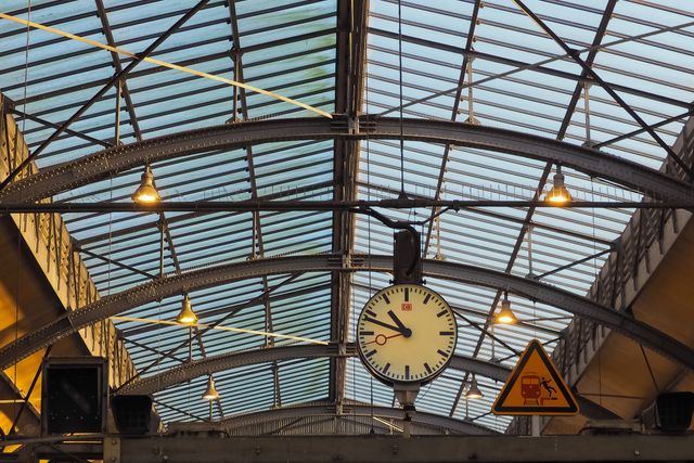 This image features an architectural glass ceiling at a train station, showcasing the blend of historic and modern design. The classic clock and iron beams indicate the intersection of timeless architecture and modern functionality. Ideal for use in articles about travel, public transport, architectural design, and urban structures.