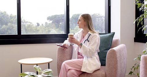 Clinic nurse taking coffee break, looking out window, holding smartphone in modern lounge