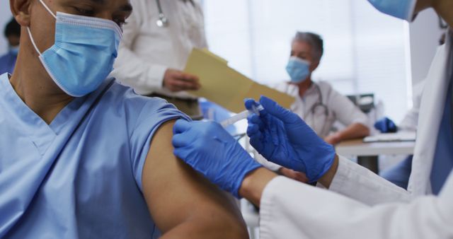 A healthcare worker is receiving a vaccine injection from a medical professional wearing gloves. The scene is set in a medical clinic where other medical staff are present. This image is useful for topics related to healthcare, vaccination campaigns, immunization programs, medical practices, and pandemic response.