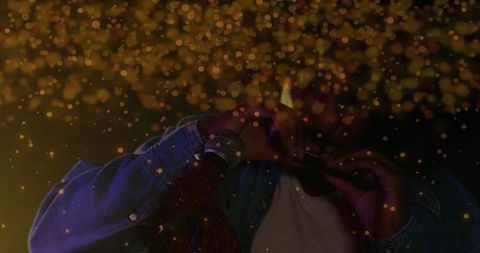 Musician in denim jacket blowing harmonica on dimly lit stage with golden bokeh