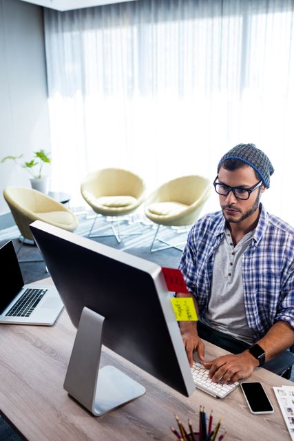 Young hipster man wearing glasses and a beanie, working at a computer desk in a modern studio. Ideal for use in articles or advertisements related to modern workspaces, creative professions, technology, and office environments.