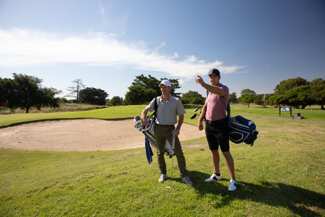 Two Caucasian male golfers practicing on a golf course on a sunny day wearing caps and golf clothes, carrying golf bags and pointing. Hobby healthy lifestyle leisure.