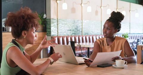African american women discussing business documents and laptop in modern cafe workspace