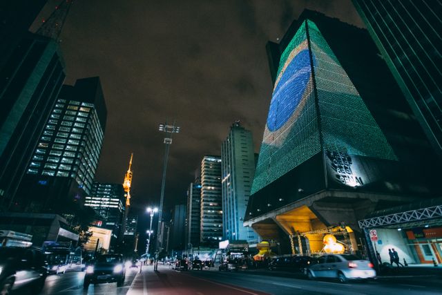 Shows the Brazilian flag illuminated on a modern skyscraper at night in Sao Paulo. Cityscape illuminated with lights from buildings and cars creates a vibrant, dynamic urban scene. Ideal for themes related to Brazilian culture, urban living, nightlife, travel, tourism and events in Sao Paulo.