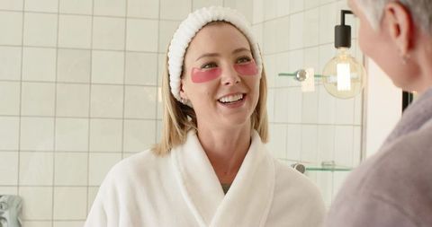 Midlife woman wearing pink under-eye patches smiling and chatting in bathroom