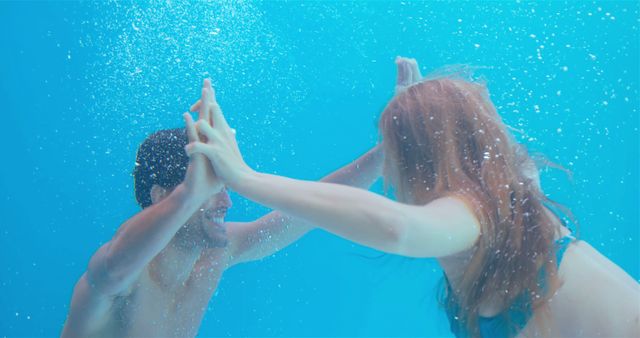 Happy couple giving each other a high-five while underwater in a swimming pool, conveying joy, teamwork, and connection. Ideal for illustrating themes of companionship, leisure activities, aquatic sports, vacation fun, relationship goals, and physical fitness.