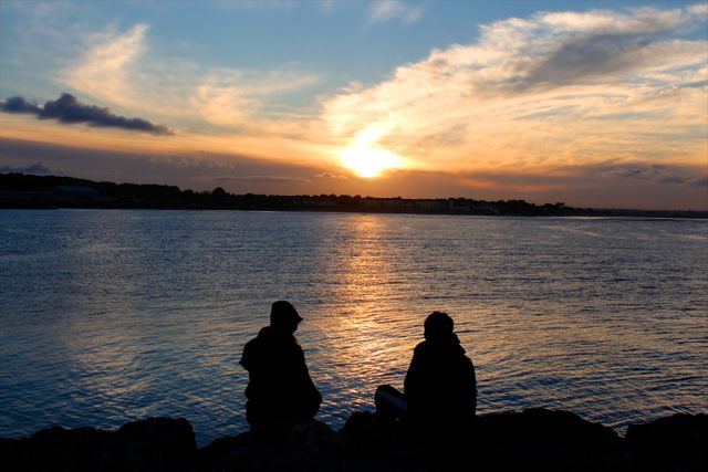 Two friends are silhouetted while sitting on the lakeshore, watching the beautiful sunset. The scene conveys a sense of peace and tranquility, perfect for themes of friendship, bonding, relaxation, and nature. Ideal for use in travel magazines, mental wellness articles, or promotional material for outdoor activities or tourism.