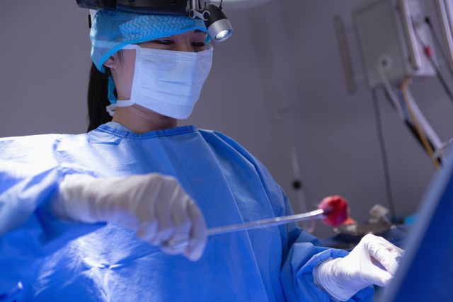 Female surgeon in blue surgical gown and mask performing an operation with focus and precision in a hospital operating room. Useful for content related to medical procedures, healthcare professions, hospital environments, and surgical practices.