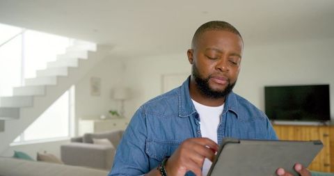 Man tapping tablet on couch in modern minimalist living room with staircase and daylight