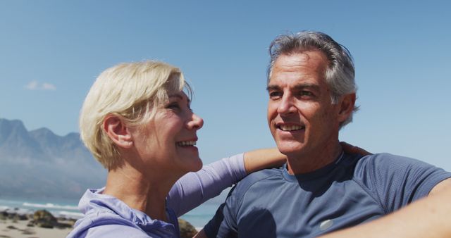 Happy mature couple embracing on beach with mountains and ocean in background. Ideal for use in advertisements or promotional material focusing on healthy lifestyles, love, wellness retreats, vacations, and senior activities. Highlights themes of happiness, togetherness, and active older adults.