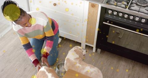 Kneeling woman feeding two dogs in cozy modern kitchen wearing colorful striped sweater