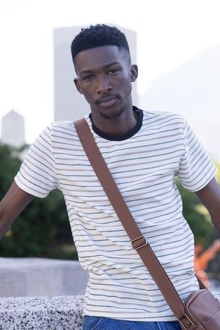 Young man leaning on concrete ledge wearing striped shirt and crossbody satchel in city plaza