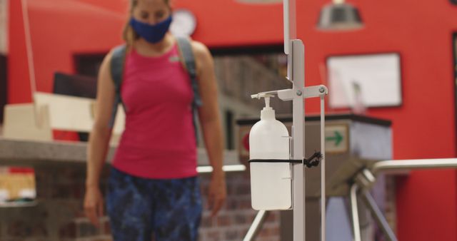 Person wearing a face mask and carrying a backpack, presumably post-workout, approaching a gym hand sanitizer station. This image can be used for articles or advertisements related to health and fitness, gym safety measures, sanitary practices during COVID-19, or general promotion of hygiene in public places.