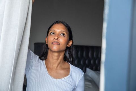 Woman contemplatively looking out bedroom window with sheer curtains