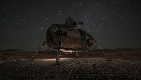Retro-futuristic landing module perched on metal grid platform under star-filled desert night sky