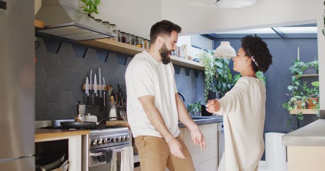 This image captures a joyful couple dancing and laughing in a modern kitchen. The bright and well-lit space features contemporary design elements, plants, and kitchen utensils, reflecting a happy home environment. Ideal for use in lifestyle blogs, relationship guides, home decor websites, and advertisements promoting happy home atmospheres or modern kitchen designs.