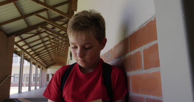 A young boy in a red shirt with a backpack is reading a book while leaning against a brick wall in a school corridor. The image conveys focus and a love for reading. Suitable for educational materials, school brochures, or articles about childhood education and literacy.