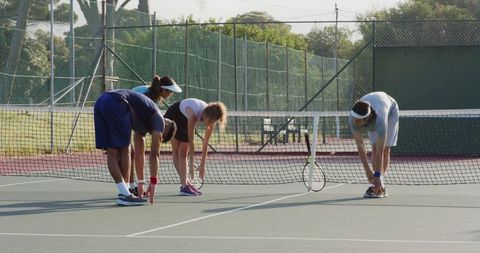 Mixed group stretching along tennis net on outdoor hard court during sunny pregame warmup