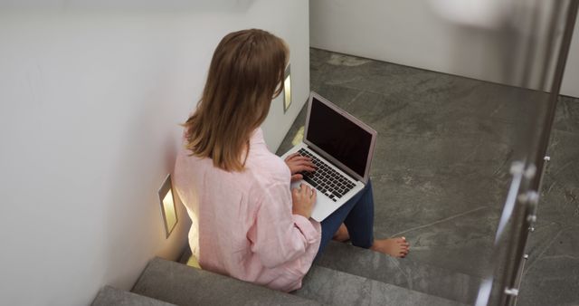 Woman working on laptop while sitting on indoor stairs. Perfect for illustrating remote work, home office environments, and modern lifestyles. Ideal for articles, blogs, or advertisements related to technology, productivity, and casual work settings.