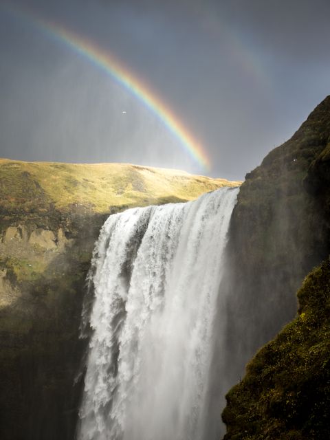 Majestic waterfall cascading over a cliff accompanied by a rainbow arching across the sky, creating a breathtaking natural spectacle. Ideal for promoting travel to Iceland, environmental awareness, nature-centric blogs, scenic poster prints, or articles on natural wonders.