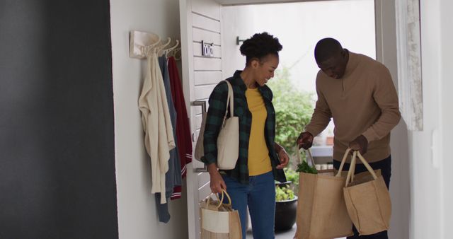 Couple entering home carrying eco-friendly reusable grocery bags. The man is carrying two brown bags filled with fresh greens. The woman has a smaller beige bag and a backpack. They appear to be unpacking groceries for meal preparation. Useful for content related to sustainable living, healthy eating, relationships, and home life.