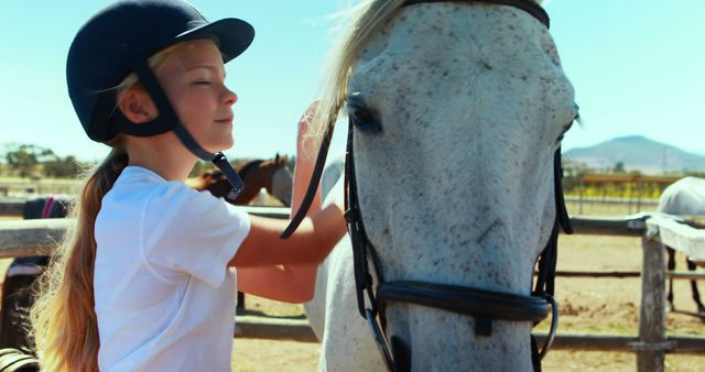 This depicts a young girl riding a white horse while smiling. She is wearing a protective riding helmet and a white shirt. The sunny day creates a vibrant environment in a countryside setting with other horses visible in the background. Ideal for illustrating themes related to youth, horsemanship, outdoor activities, farm life, and summertime adventures.