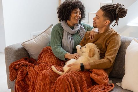 Couple cuddling with puppy on sofa under rust blanket, sharing cozy at-home moment