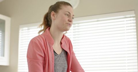 Young woman standing by bright kitchen window blinds wearing coral jacket looking contemplative