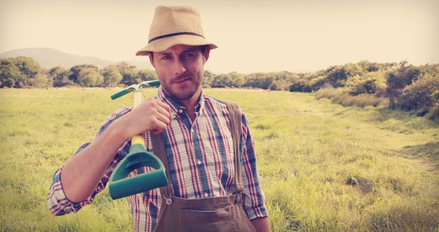A farmer wearing a straw hat, plaid shirt, and overalls is holding a garden tool over his shoulder in a grassy field. This can be used to depict themes related to agriculture, rural lifestyle, manual labor, and farming. Perfect for advertisements, blogs, and articles focusing on farming, country living, and nature.