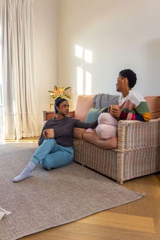 Diverse women sharing tea and chatting on wicker sofa in sunlit minimalist living room