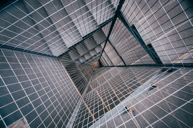 Unique perspective looking down through industrial metal grated staircase in modern building. Geometric patterns and textures create abstract and minimalist feel. Suitable for use in architectural design projects, abstract art collections, and promotional materials emphasizing modern or industrial design.