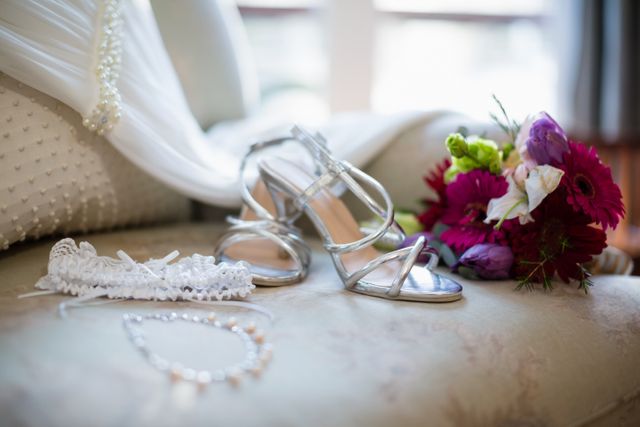 Elegant wedding accessories including silver shoes, a floral bouquet, a lace garter, and jewelry arranged on a sofa. Perfect for use in wedding planning materials, bridal magazines, and romantic event promotions.