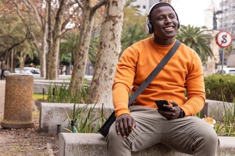 Young man listening to music on bench in urban park holding smartphone and snack