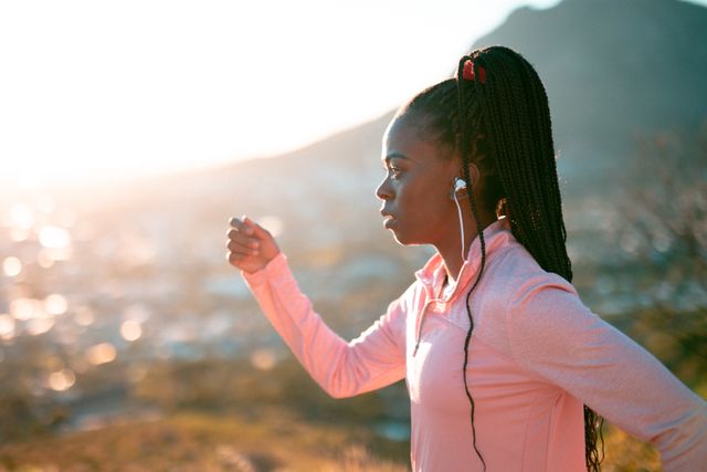 African American woman jogging in countryside during sunrise, wearing wireless earphones and sportswear. Ideal for promoting healthy lifestyle, outdoor fitness, and athletic motivation. Suitable for fitness blogs, health magazines, and sportswear advertisements.