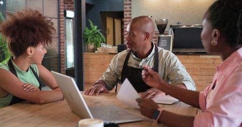 African american cafe team collaborating and planning over laptop and papers in rustic coffee shop