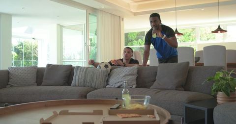 Friends watching soccer match holding soccer ball and drinking from bottle in bright living room