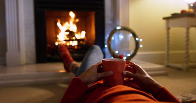 Person lounging by a fireplace, enjoying a hot beverage in a cozy home interior. Perfect for themes involving winter holidays, relaxation, comfort, home, and hygge. Great for use in articles and advertisements about home comfort, holiday getaway, and cozy lifestyle.