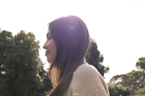 Asian woman standing in sunlit park with backlit hair, wearing beige coat and serene profile