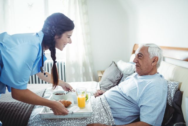 Nurse serving breakfast to senior man in bed, showcasing home healthcare and caregiving. Ideal for use in articles or advertisements about elderly care, home nursing services, and healthcare support. Highlights the importance of professional assistance and comfort for seniors.