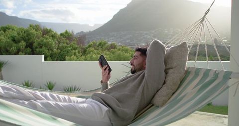 Indian man relaxing in striped hammock on sunlit terrace with smartphone and mountain view