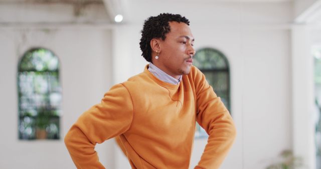 This image shows a confident young man with curly hair wearing an orange sweater and standing indoors. The modern and stylish ambiance makes it versatile for use in fashion blogs, lifestyle magazines, or promotional material for casual or stylish clothing.