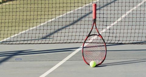 Red and Black Tennis Racket Leaning Against Net with Yellow Ball on Hard Court