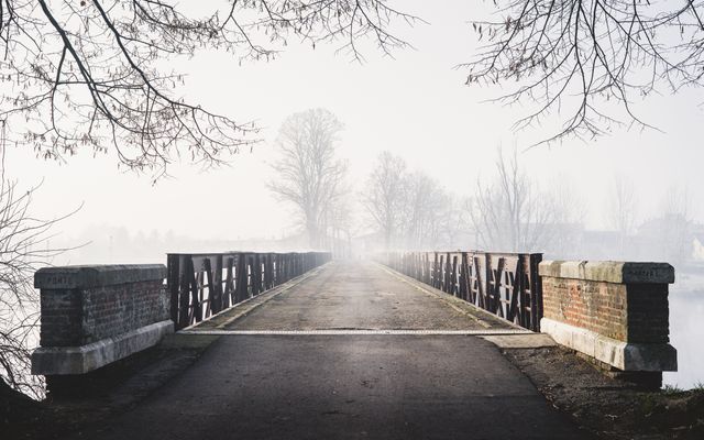 Foggy morning on historic stone bridge with empty walkway extending into misty background. Bare trees arching over and tranquil river setting creates tranquil and calm atmosphere. Ideal for themes related to nature, peace, serenity, solitude, or picturesque landscapes.