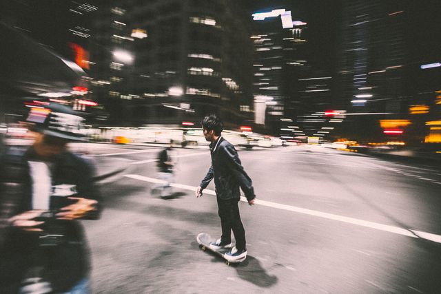 Young man skateboarding on busy city street at night with blurred lights and surroundings. Ideal for themes of urban lifestyle, youth culture, night activities, and dynamic city life. Can be used in advertisements for sports equipment, urban lifestyle campaigns, and visual storytelling involving modern city living.