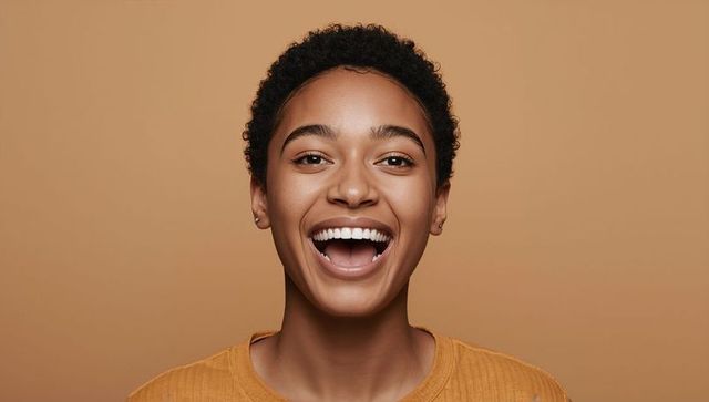 Joyful young woman laughing with short curly hair wearing mustard top warm tan background