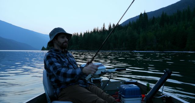 Photo captures a man fishing on a calm mountain lake during sunset, wearing a plaid shirt and hat. Ideal for promoting outdoor activities, fishing gear, travel destinations, relaxation, and nature excursions.