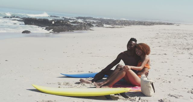 Happy biracial couple sitting with surfboards on sunny beach embracing. Summer, hobbies, free time, surfing, romance and vacations.