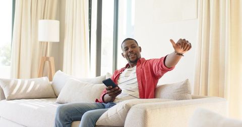 African american man relaxing on sofa holding smartphone and giving thumbs-up gesture