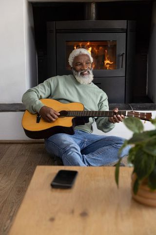 Senior african american man playing acoustic guitar by wood stove with cozy firelight